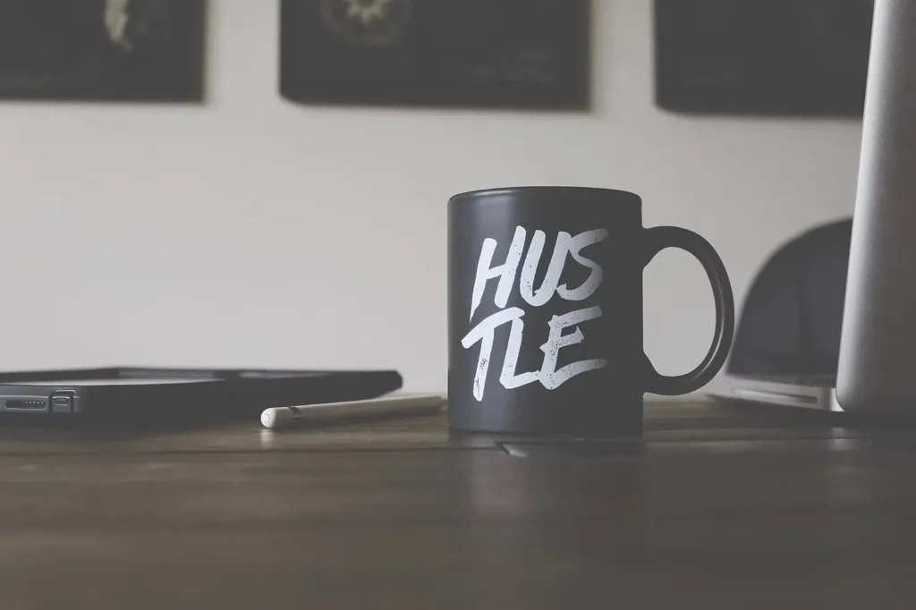 Hustle mug beside a laptop and tablet representing workplace pressure and burnout