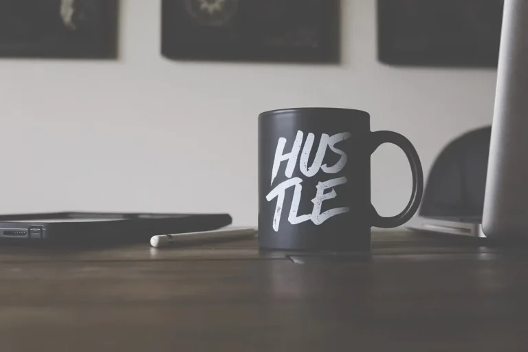 Hustle mug beside a laptop and tablet representing workplace pressure and burnout